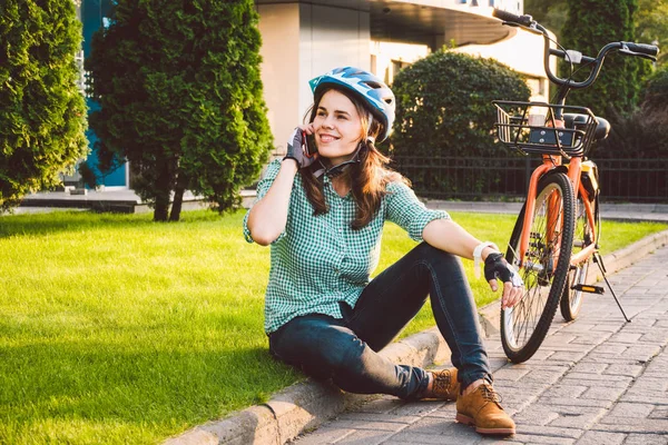 İnsan ve şehir bisikleti, çevre dostu ulaşım. Çimlerin üzerinde dinlenen güzel beyaz kadın işçi turuncu bisikletli kırmızı bir cep telefonu kullanıyor. — Stok fotoğraf