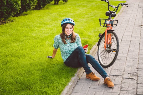 İnsan ve şehir bisikleti, çevre dostu ulaşım. Çimlerin üzerinde dinlenen güzel beyaz kadın işçi turuncu bisikletli kırmızı bir cep telefonu kullanıyor. — Stok fotoğraf