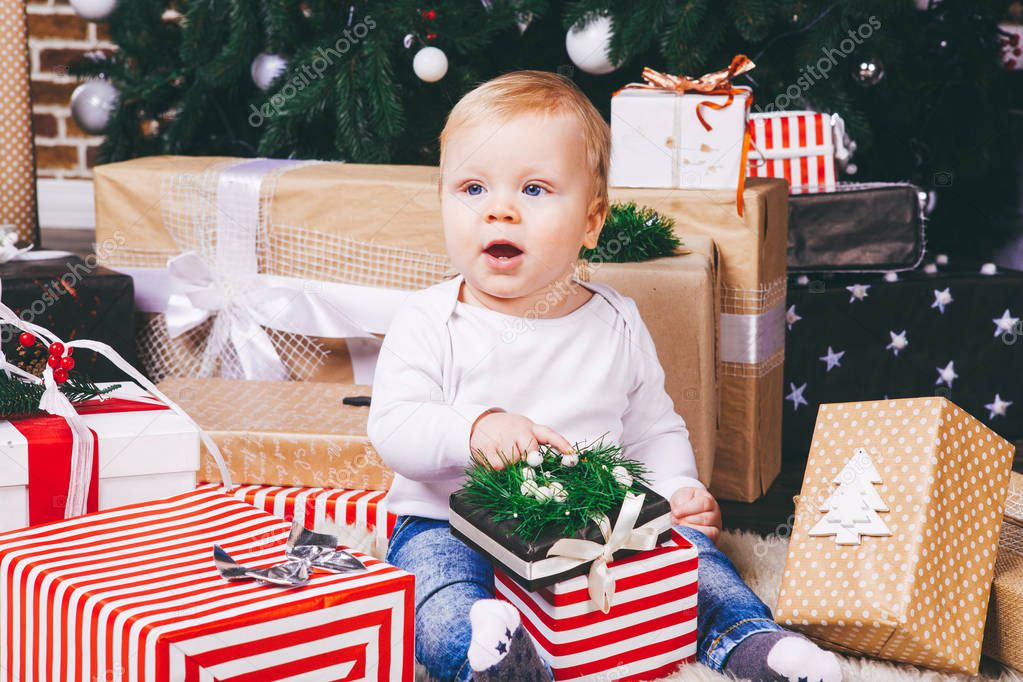 theme winter and Christmas holidays. Child boy Caucasian blond 1 year old sitting home floor near Christmas tree with New Year decor on shaggy carpet skin receives gifts, opens gift boxes in evening.