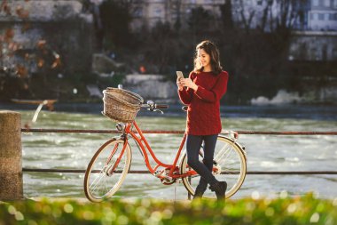 Tema Turizm bisiklet ve modern teknoloji. Güzel genç kafkas kadın standları nehir Nehri Rhine Basel İsviçre kış sıcak güneşli hava üzerinde kırmızı retro Bisiklet yakınındaki elinde telefon kullanır.