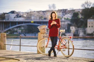 Tema Turizm bisiklet ve modern teknoloji. Güzel genç kafkas kadın standları nehir Nehri Rhine Basel İsviçre kış sıcak güneşli hava üzerinde kırmızı retro Bisiklet yakınındaki elinde telefon kullanır.