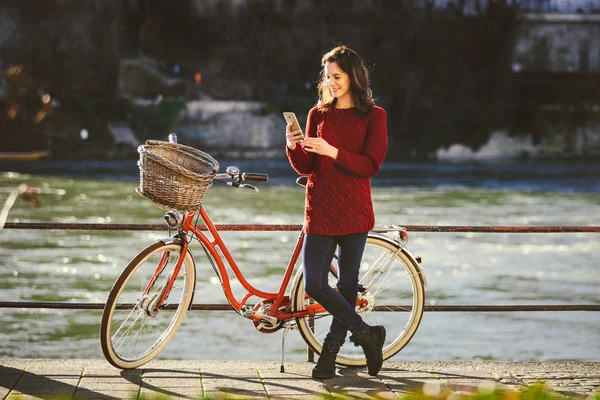 theme tourism on bicycle and modern technology. Beautiful young caucasian woman stands near red retro bicycle on riverside river Rhine Basel Swiss winter warm sunny weather uses the phone in hand.