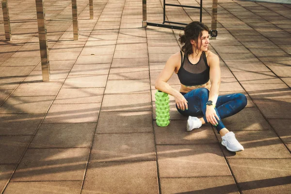 theme sport and rehabilitation sports medicine. Beautiful strong slender Caucasian woman athlete sits next foam roller green field street workout to remove the pain, stretch and massage muscles.