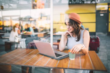 Bir kafede bir terasta yaz şapka ve bir tabak içinde ahşap bir masada oturan güzel genç beyaz köpek yavrusu bir dizüstü rezervasyon otel gezi yaz aylarında çalışma teknolojisini kullanır.