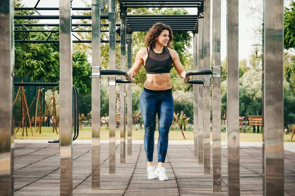 The theme women sports and health. Beautiful sexy caucasian woman with curly long hair posing on outdoor sports ground holding horizontal bar with tattoo on stomach in sports top and tight pants.