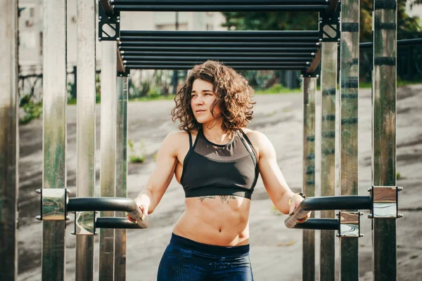 O tema mulheres esportes e saúde. Mulher caucasiana sexy bonita com cabelos longos encaracolados posando no chão de esportes ao ar livre segurando barra horizontal com tatuagem no estômago em esportes top e calças apertadas — Fotografia de Stock