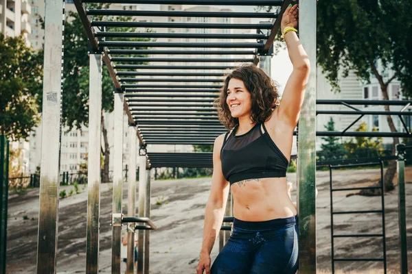 The theme women sports and health. Beautiful sexy caucasian woman with curly long hair posing on outdoor sports ground holding horizontal bar with tattoo on stomach in sports top and tight pants