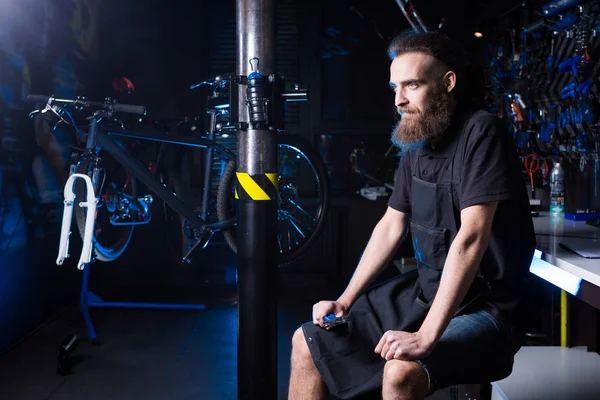 Küçük işletme sahibi sakallı genç adamın portresi. Bisiklet dükkanı arka planda apronda çalışma siyah giysili elinde aracıyla oturan adam Bisiklet mekanik atölye işçisi.