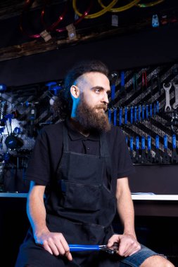 Küçük işletme sahibi sakallı genç adamın portresi. Bisiklet dükkanı arka planda apronda çalışma siyah giysili elinde aracıyla oturan adam Bisiklet mekanik atölye işçisi.