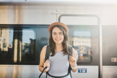 Tema demiryolu ve seyahat. Dişlek ile genç beyaz kadın portre gülümseme, sırt çantası ve Turizm izole mat elbise ve şapka yaz aylarında araçlar ile Tren İstasyonu Tren arka plan ayakta.