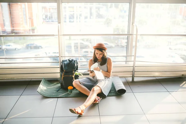 Waiting, delayed transport in the terminal of the airport or train station. Young caucasian woman in dress and hat sits on tourist rug with backpack near window to room and looks into tourist map..