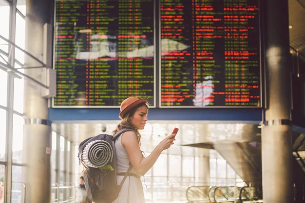 Tema seyahat ve tranosport. Güzel genç kafkas kadın elbise ve tren istasyonuna veya kırmızı telefon tutarak bir zamanlama seyir terminal içinde duran sırt çantası iletişim teknolojisini kullanır.