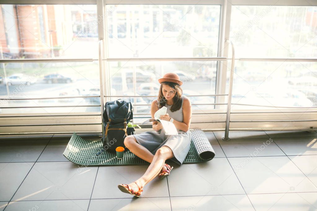 Waiting, delayed transport in the terminal of the airport or train station. Young caucasian woman in dress and hat sits on tourist rug with backpack near window to room and looks into tourist map..