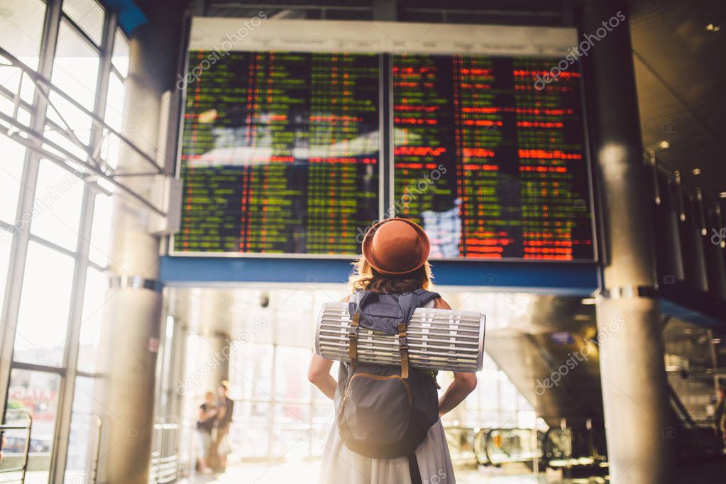 Theme travel public transport. young woman standing with back in dress and hat behind backpack and camping equipment for sleeping, insulating mat looks schedule on scoreboard airport station sunny day