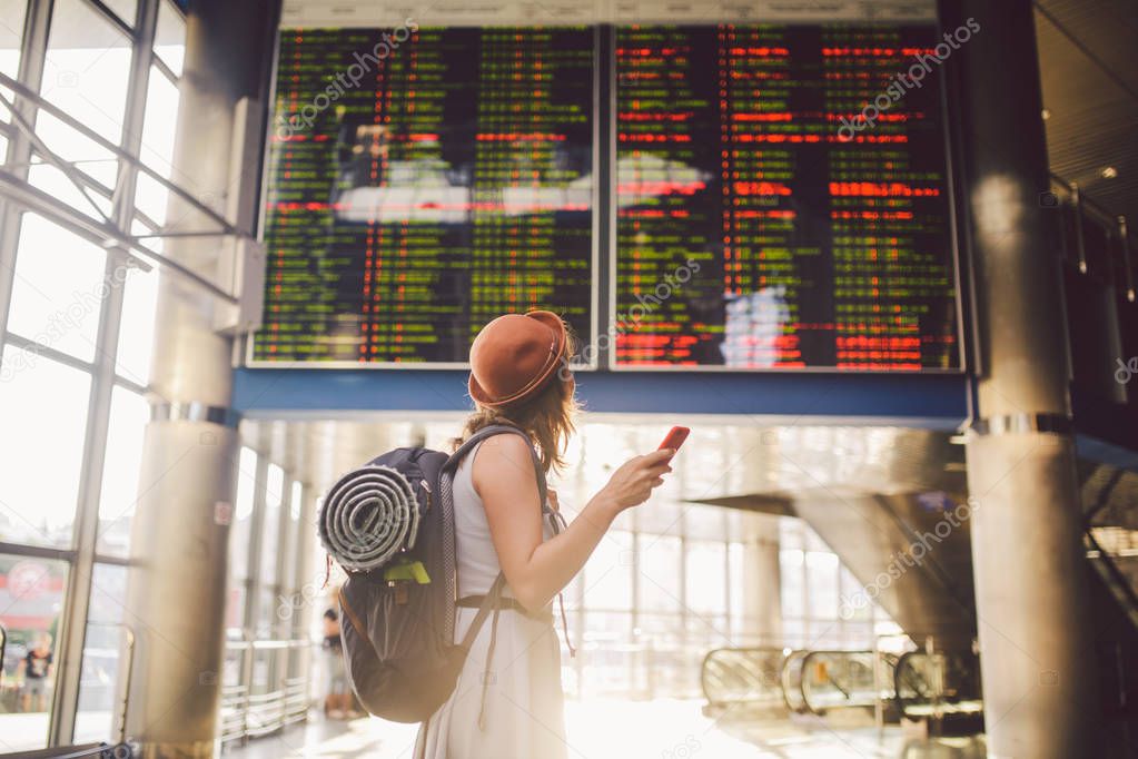 Theme travel and tranosport. Beautiful young caucasian woman in dress and backpack standing inside train station or terminal looking at a schedule holding a red phone, uses communication technology.