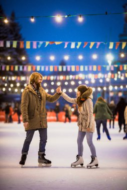 Uzun saçlı sarışın saçlı beyaz adam genç çift tr ve eğlenceli, aktif tarihi buz pateni Buz arenada akşam şehir meydanında Noel arifesinde kışın sakal ve güzel bir kadın var