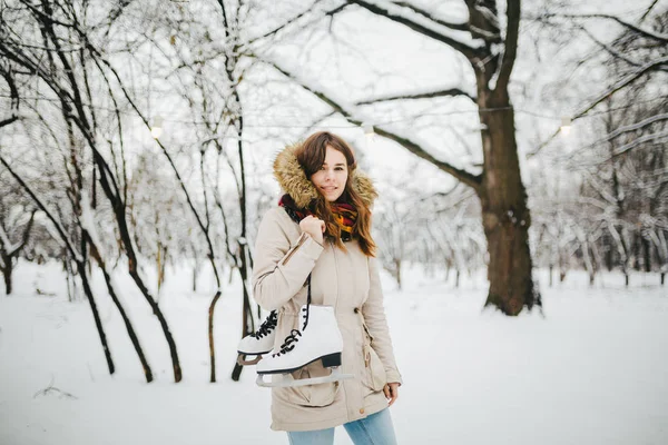 Theme is weekend holiday in winter. A beautiful young Caucasian woman stands in a snow covered park in jacket with hood and fur in jeans and holds on shoulder pair of white skates for long shoelaces.