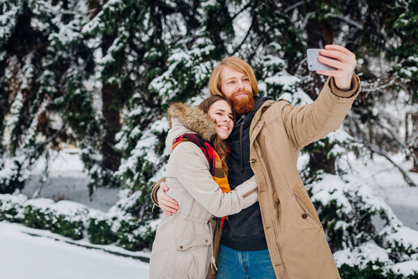 Young couple in love make joint photo of themselves on mobile phone. The guy holds smartphone on his elongated hand and makes selfie photo on background of park with coniferous trees in winter.