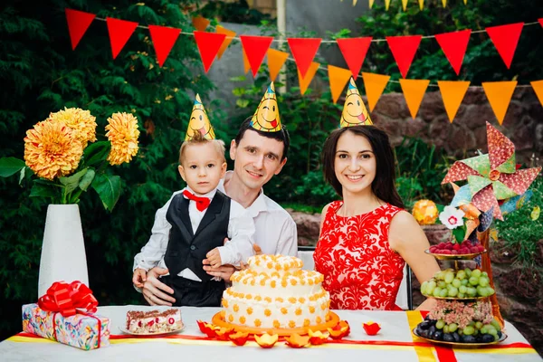 Barn födelsedag tema. familj med tre kaukasiska personer sitter i bakgården av huset på ett festligt dukade bordet i roliga hattar och mössor på huvudet. Glädje och lycka kaka och bakelse mellanmål — Stockfoto