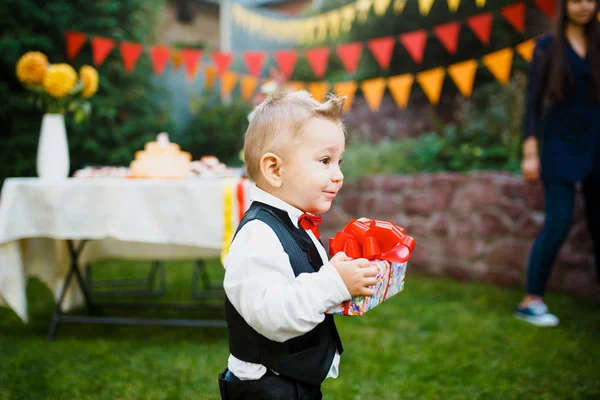 誕生日の驚き。少年は、ケーキでお祝いテーブルの背景に庭のギフトを持つボックスを保持しています。黒いスーツと赤い蝶に身を包んだ — ストック写真