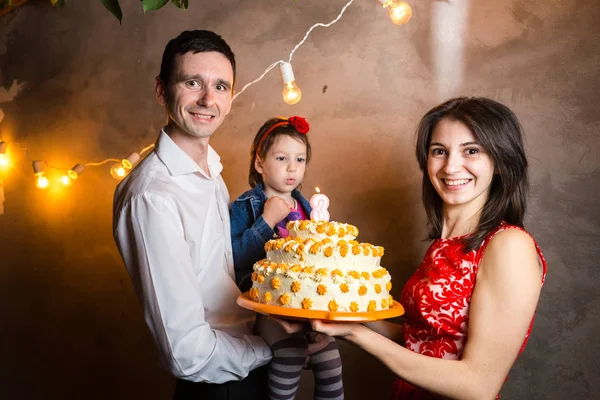Theme family holiday childrens birthday and blowing out candles on large cake. young family of three people standing and holding 5 year old daughter in yard against gray wall and garland yellow bulbs