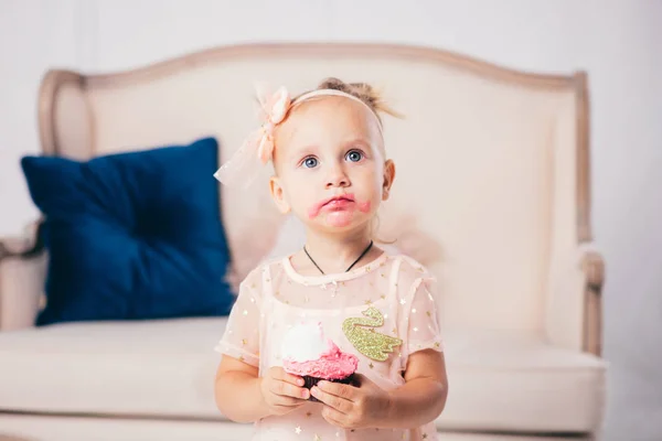 stock image childrens birthday. funny two-year-old Caucasian girl in pink dress standing to bedroom of house the background of couch and eating, holding cake dessert. Face and mouth smeared, smeared with cream