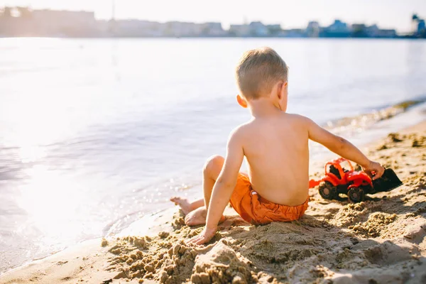 Enfant caucasien garçon jouant jouet tracteur rouge, pelle sur une plage de sable près de la rivière en short rouge au coucher du soleil jour — Photo