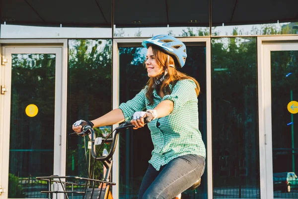 Theme to work on the bike. A young Caucasian woman arrived on environmentally friendly transport bike to the office. Girl in a bicycle parking office building in a helmet, gloves and shirt and jeans.