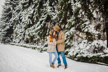 Genç beyaz erkek ve kız aşk heteroseksüel çift açık havada bir kucaklama bir karlı kozalaklı ayakta arka plan üzerinde bir kış parkta buluşacağım. Adam kucaklama kadın Sevgililer günü