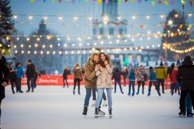 Uzun saçlı sarışın saçlı beyaz adam genç çift tr ve eğlenceli, aktif tarihi buz pateni Buz arenada akşam şehir meydanında Noel arifesinde kışın sakal ve güzel bir kadın var