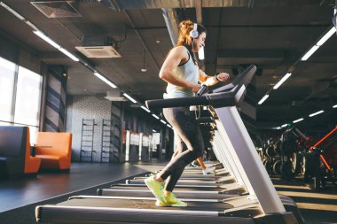 Güzel genç kadın uzun saçlı tren bir koşu bandı üzerinde spor salonunda. Büyük kulaklıklar müzik dinler. Başına bir spor saat hızı hızı, kalp hızı, izleme ve kronometre için siyah kol.