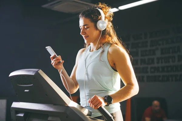 Theme sport and music. A beautiful Caucasian woman running in gym on treadmill. On head big white headphones, the girl listens to music during a cardio workout for weight loss and uses phone in hand.