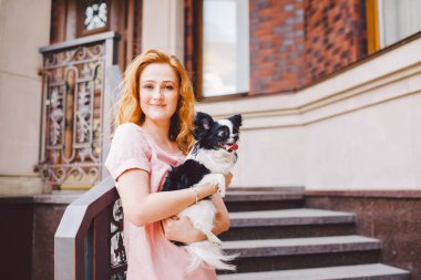 Kırmızı uzun saçlı güzel bir genç kadın bir küçük, şirin komik büyük gözlü köpek iki çiçek, siyah-beyaz evde beslenen hayvan hichuahua karşı bir ev yaz aylarında kırmızı tuğla nesil tutuyor.