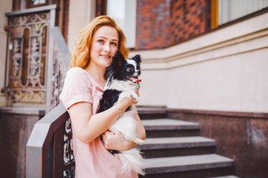 Kırmızı uzun saçlı güzel bir genç kadın bir küçük, şirin komik büyük gözlü köpek iki çiçek, siyah-beyaz evde beslenen hayvan hichuahua karşı bir ev yaz aylarında kırmızı tuğla nesil tutuyor.