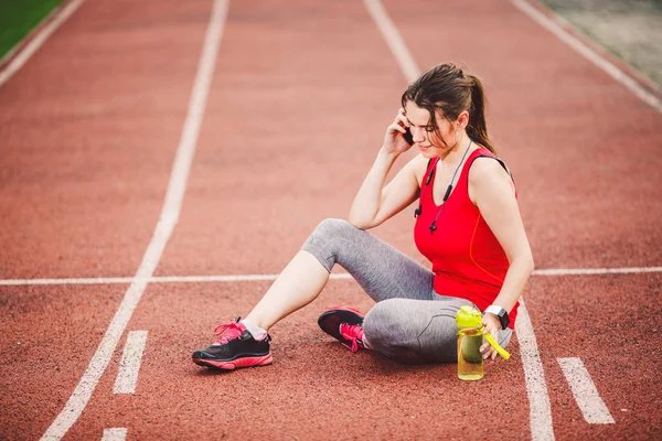 体育和技术 一个美丽的年轻的白种女人与马尾辫坐在休息后 在体育场跑步运动期间 一个红色的跑步机轨道 用于手机耳机和运动手表 — 图库照片