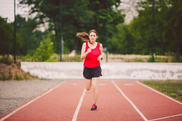 Beautiful young athlete Caucasian woman with big breasts in red T-shirt and short shorts running jog, running in a stadium with a red rubber coating. Hairstyle of ponytail