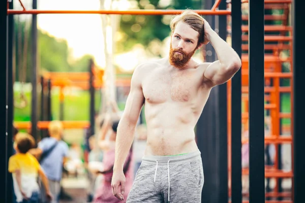 Subject sport street workout Handsome young caucasian man with a bare-chested muscular with long red hair and beard posing outdoor sports ground, gym outdoors. Sexy guy touches his hair
