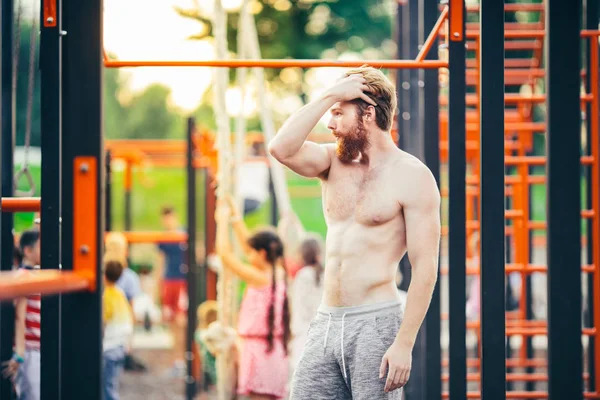 Subject sport street workout Handsome young caucasian man with a bare-chested muscular with long red hair and beard posing outdoor sports ground, gym outdoors. Sexy guy touches his hair