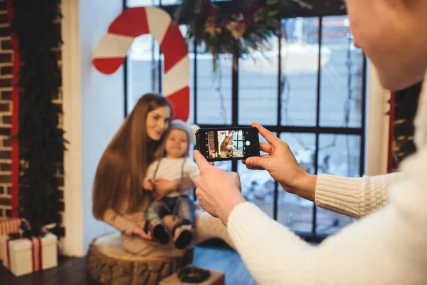 Theme mobile photography, amateur photo and video on phone. Hands Caucasian man holds uses smartphone makes photo of mother and son at home near window in winter in Christmas time New Years decor.