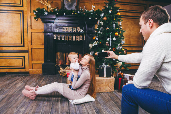 Theme mobile photography, amateur photo video phone. Hands Caucasian man holds uses smartphone makes photo mother and son home near fireplace and Christmas tree winter Christmas time New Year decor.