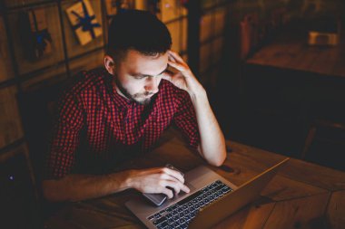 Genç yakışıklı bir beyaz adam bir sakal ve ahşap bir masa, bir dizüstü bilgisayar yanında oturan kırmızı Kareli gömlekli dişlek gülümseme ile portresi. Akşamları coffee shop