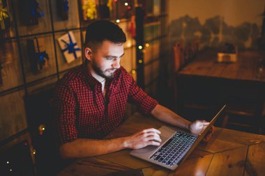 Genç yakışıklı bir beyaz adam bir sakal ve ahşap bir masa, bir dizüstü bilgisayar yanında oturan kırmızı Kareli gömlekli dişlek gülümseme ile portresi. Akşamları coffee shop