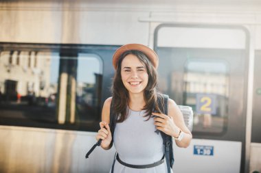 Tema demiryolu ve seyahat. Dişlek ile genç beyaz kadın portre gülümseme, sırt çantası ve Turizm izole mat elbise ve şapka yaz aylarında araçlar ile Tren İstasyonu Tren arka plan ayakta.