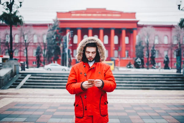 A handsome young guy with beard and red jacket in hood a student uses a mobile phone, writes, writes a correspondence over the phone with a smile against the red building of the university or college.