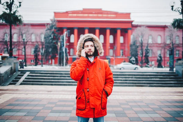 A handsome young guy with beard and red jacket in hood a student uses a mobile phone, writes, writes a correspondence over the phone with a smile against the red building of the university or college.