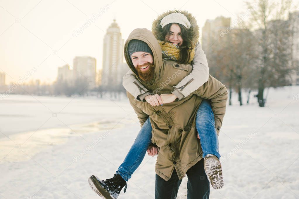 Theme New Year Christmas Mood Winter Snow Holidays Valentine Day. Young Caucasian couple lovers joy, laughter fooling water in city park. man holds woman on shoulders as backpack.