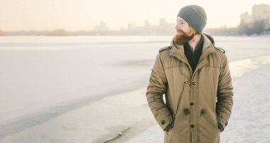 Yakın çekim beyaz genç erkek kırmızı saç ve sakal bir şapka ve kış modeli kar dondurmak için bir göl çerçevede bir poz bir park kat.