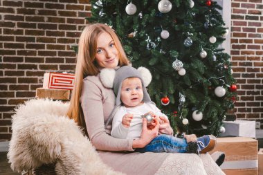 Tema Noel tatil kış yeni yıl. Genç bir şık beyaz anne oğlunu kucağında bir kızak üzerinde oturan ve bir şekilde dekore edilmiş ev ağacın altında kürk giymiş komik gömlekli 1 yıl için tutar