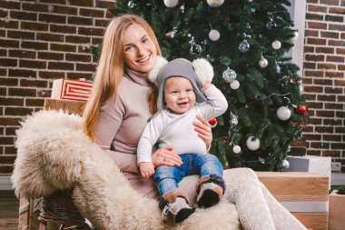 Tema Noel tatil kış yeni yıl. Genç bir şık beyaz anne oğlunu kucağında bir kızak üzerinde oturan ve bir şekilde dekore edilmiş ev ağacın altında kürk giymiş komik gömlekli 1 yıl için tutar