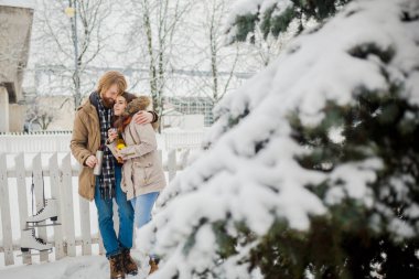 Genç Çift sarı uzun saçları ve sakalı beyaz adamla sevgi dolu, güzel bir kadın iyi eğlenceler termos sıcak bir içecek içme beyaz çit ve iğne yapraklı ağaç kar parkı yeşil elma yeme.
